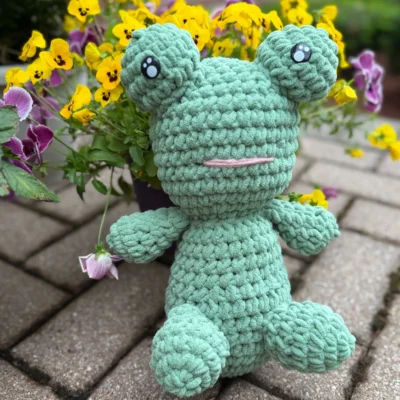 green frog crochet plushie sitting by yellow and purple flowers